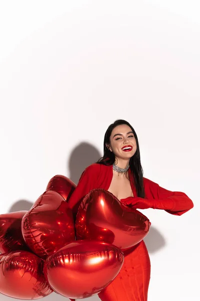 Stilvolle Frau mit roten Lippen blickt in die Kamera in der Nähe herzförmige Luftballons auf weißem Hintergrund mit Schatten — Stockfoto