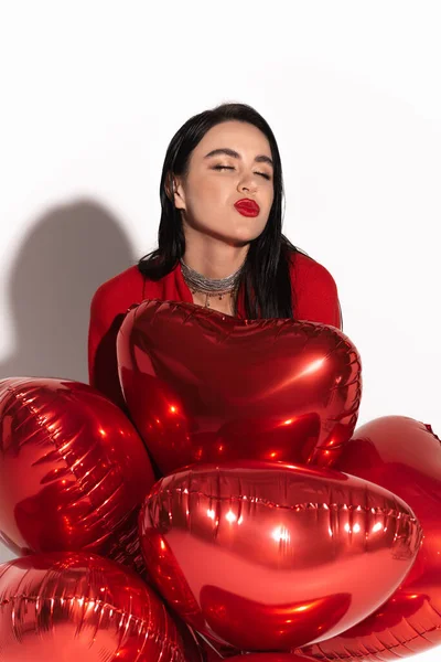 Elegante donna con labbra rosse che soffiano bacio vicino a palloncini a forma di cuore su sfondo bianco con ombra — Foto stock
