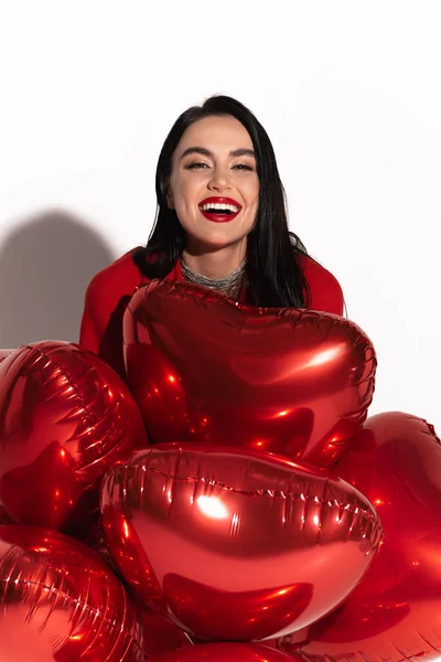 Modelo morena positivo com lábios vermelhos olhando para a câmera perto de balões em forma de coração no fundo branco com sombra — Fotografia de Stock