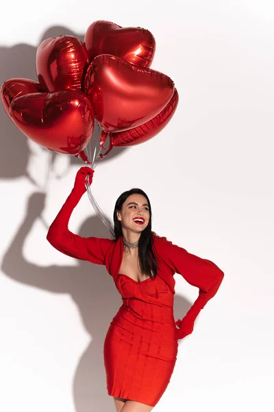 Trendy Frau im roten Kleid hält herzförmige Luftballons auf weißem Hintergrund mit Schatten — Stockfoto