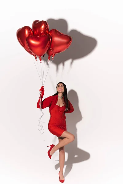 Elegante donna in abito rosso e tacchi guardando palloncini a forma di cuore su sfondo bianco con ombra — Foto stock