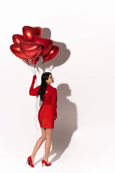 Vue latérale du modèle souriant en robe rouge et talons tenant des ballons festifs en forme de cœur sur fond blanc avec ombre — Photo de stock