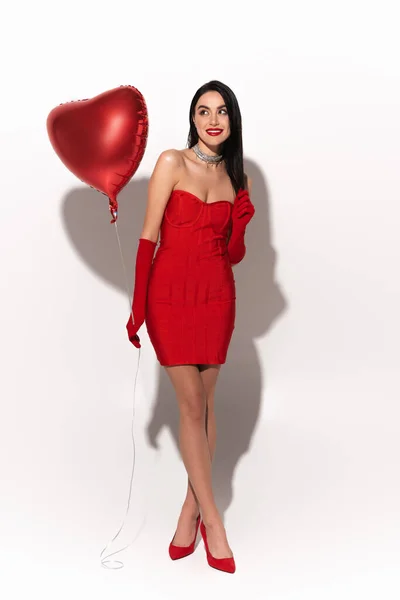 Longitud completa del modelo elegante en vestido rojo que sostiene el globo en forma de corazón sobre fondo blanco con sombra - foto de stock