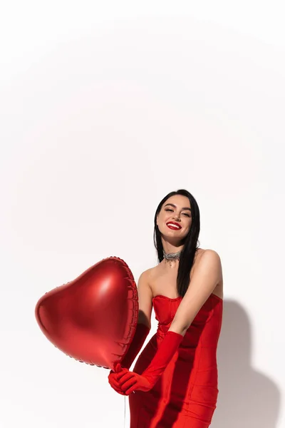 Femme souriante en robe rouge tenant des ballons en forme de coeur et regardant la caméra sur fond blanc avec ombre — Photo de stock