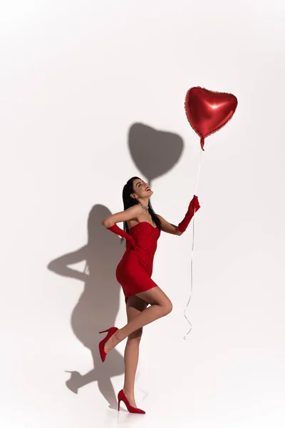 Femme élégante en talons rouges et robe posant et regardant ballon en forme de coeur sur fond blanc avec ombre — Photo de stock
