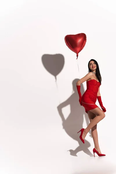 Sexy brunette woman in heels and red gloves holding heart shaped balloon on white background with shadow — Stock Photo