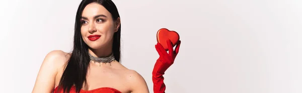 Модная брюнетка женщина с красными губами держа подарок в форме сердца на сером фоне, баннер — стоковое фото