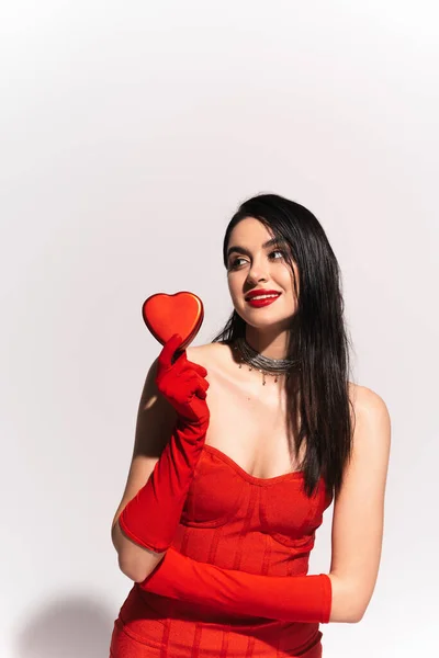 Elegante mujer en vestido rojo y guantes sosteniendo caja de regalo en forma de corazón sobre fondo gris - foto de stock