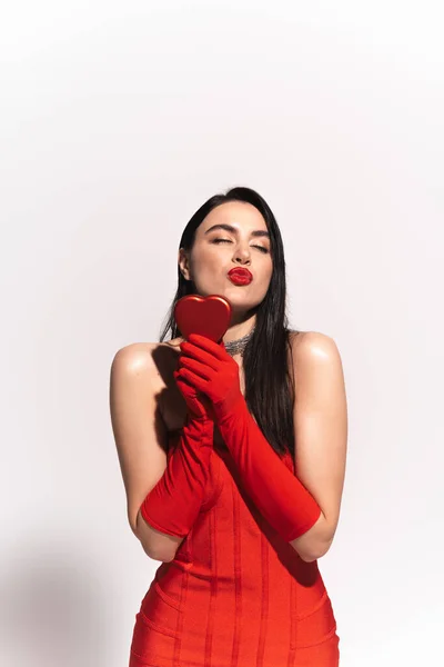 Modelo elegante en vestido rojo y guantes que sostienen la caja de regalo en forma de corazón y el beso de aire soplado sobre fondo gris - foto de stock