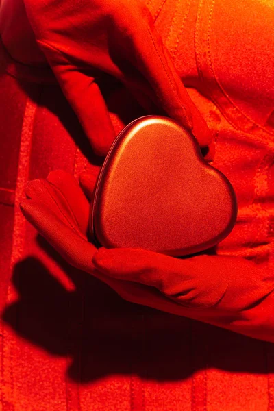 Обрізаний вид жінки в червоній сукні і рукавички, що тримають подарункову коробку у формі серця — стокове фото