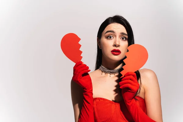 Занепокоєна жінка в червоних рукавичках тримає зламане паперове серце ізольовано на сірому — стокове фото