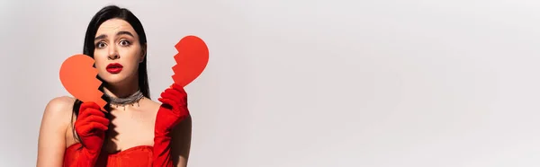 Mujer preocupada con guantes rojos sosteniendo papel roto corazón aislado en gris, bandera — Stock Photo
