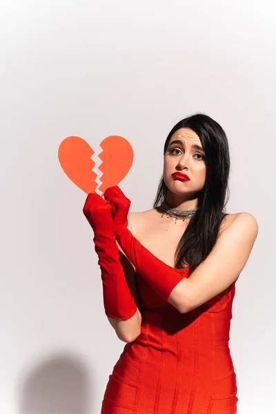 Mujer disgustada con guantes rojos y vestido sosteniendo el corazón de papel roto sobre fondo gris - foto de stock