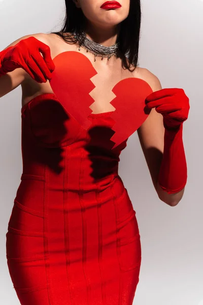 Vista ritagliata di donna elegante sconvolto in abito rosso tenendo rotto cuore di carta isolato su grigio — Foto stock
