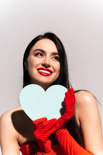 Smiling brunette woman in red gloves holding paper heart isolated on grey — Stock Photo