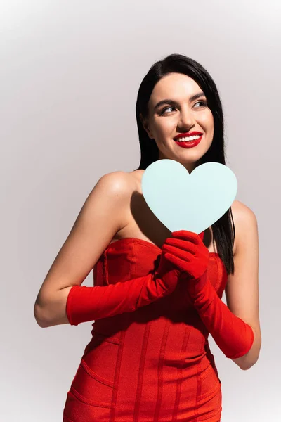Позитивная и стильная женщина в красных перчатках и платье с бумажным сердцем, изолированным на сером — стоковое фото