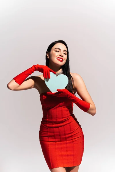 Pleased woman in red dress holding paper heart isolated on grey — Stock Photo