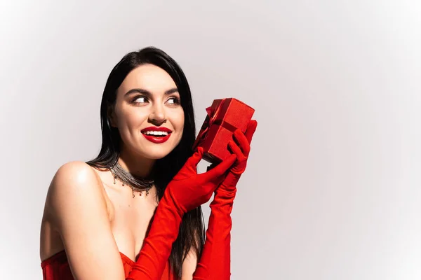 Mujer morena alegre con guantes rojos sosteniendo presente aislado en gris - foto de stock