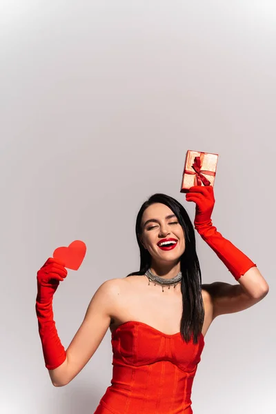 Fröhliche brünette Frau in roten Handschuhen hält Geschenk und Papierherz isoliert auf grau — Stockfoto