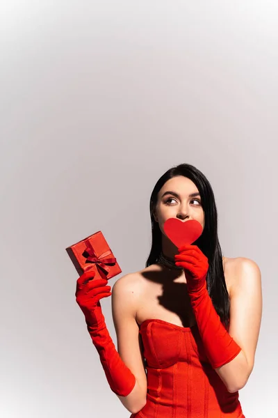Mujer morena con estilo en guantes rojos sosteniendo el corazón de papel y regalo aislado en gris - foto de stock