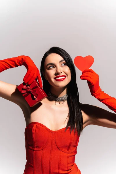Весела жінка в червоних рукавичках і сукні тримає подарункову коробку і паперове серце ізольовано на сірому — стокове фото