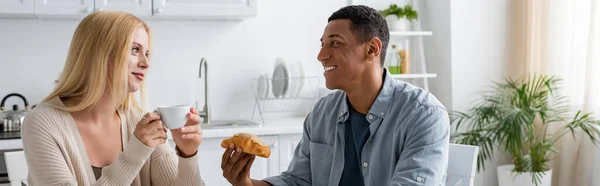 Complacida pareja multiétnica con taza de café y delicioso croissant mirándose en la cocina, pancarta - foto de stock
