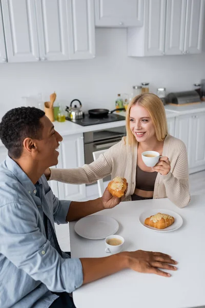 Feliz pareja interracial mirándose unos a otros cerca de croissants y café en la cocina - foto de stock