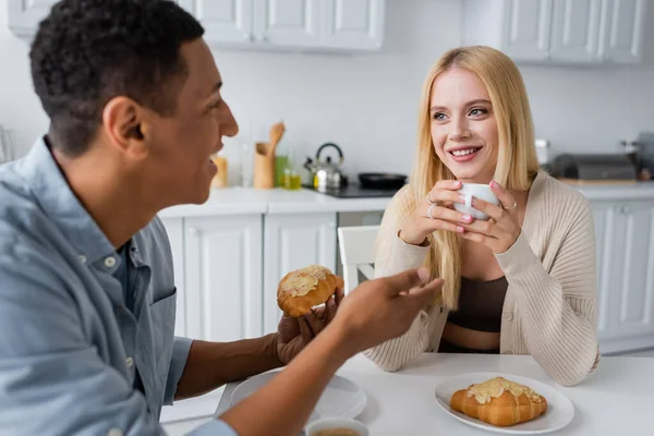Borrosa afroamericano hombre con sabroso croissant hablando con sonriente novia rubia con taza de café - foto de stock