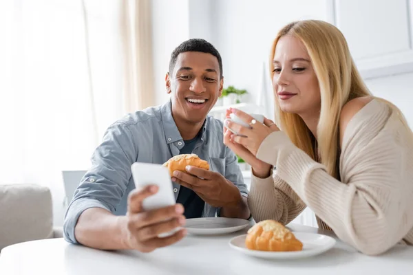 Cheerful african american man showing blurred smartphone to smiling girlfriend during breakfast in kitchen — Stock Photo