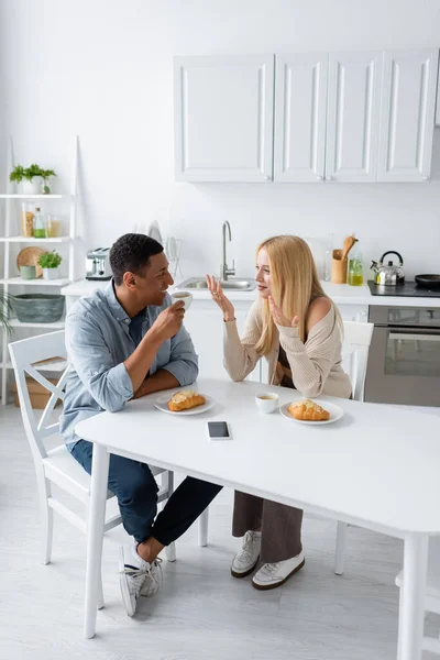Feliz pareja interracial hablando cerca de tazas de café y croissants mientras desayuna en la cocina - foto de stock