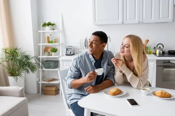 Alegre casal multiétnico olhando para longe perto de café e deliciosos croissants na cozinha — Fotografia de Stock