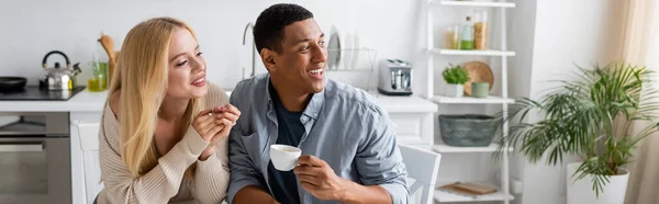 Alegre afroamericano hombre sosteniendo taza de café y mirando hacia otro lado cerca rubia novia, pancarta - foto de stock