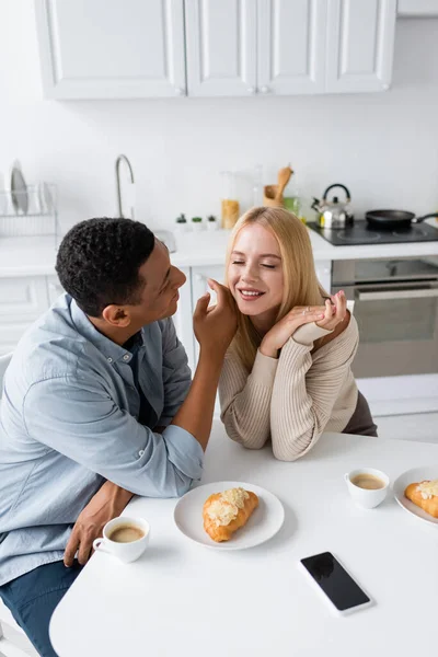 Afrikanisch-amerikanischer Mann berührt Gesicht einer hübschen blonden Freundin in der Nähe von Kaffeetassen und Croissants in der Küche — Stockfoto