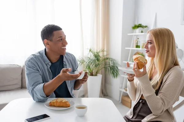 Homme afro-américain gai avec tasse de café parler à petite amie blonde avec croissant savoureux dans la cuisine — Photo de stock