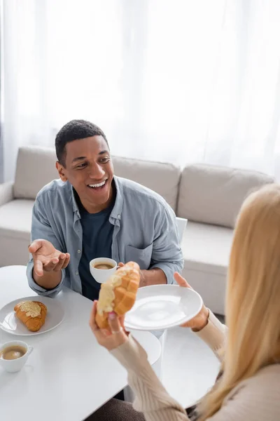 Felice uomo africano americano con tazza di caffè parlare con la ragazza bionda in possesso di gustoso croissant — Foto stock