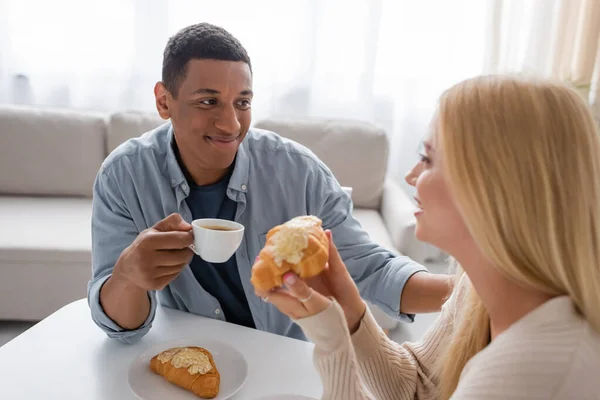 Rubia borrosa sosteniendo delicioso croissant cerca complacido hombre afroamericano con taza de café - foto de stock