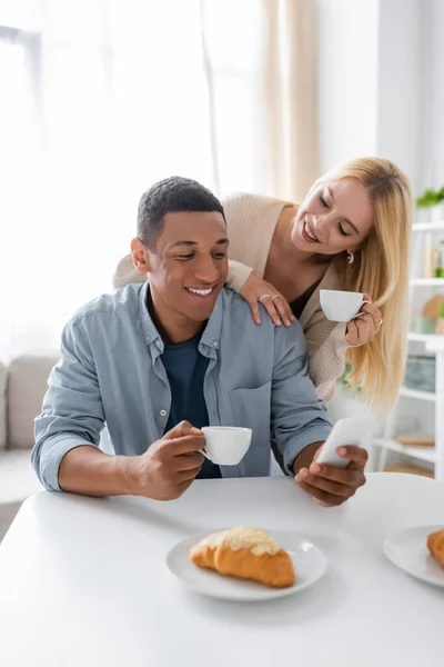 Блондинка с чашкой кофе улыбается рядом с африканским американским парнем, используя смартфон во время завтрака — стоковое фото