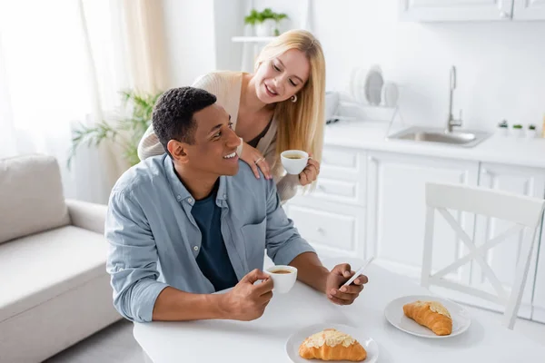 Mujer rubia sosteniendo taza de café cerca de hombre afroamericano usando teléfono inteligente durante el desayuno en la cocina - foto de stock