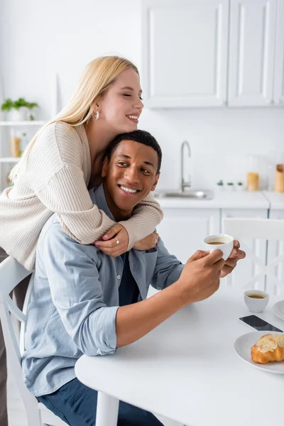 Felice donna bionda ad occhi chiusi che abbraccia il fidanzato africano americano con tazza di caffè — Foto stock