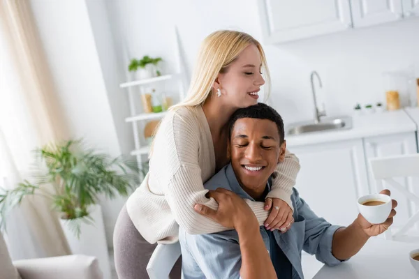 Mulher loira feliz abraçando o namorado americano africano sorrindo com os olhos fechados e segurando xícara de café — Fotografia de Stock