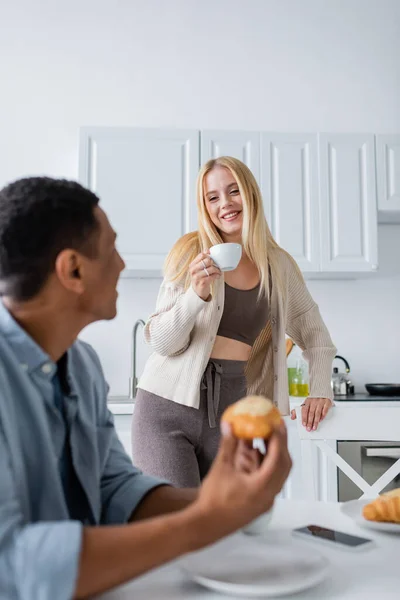Hübsche blonde Frau mit Kaffeetasse, die verschwommenen afrikanisch-amerikanischen Freund beim Frühstück in der Küche betrachtet — Stockfoto