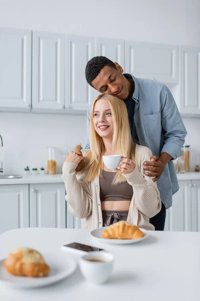 Africano americano uomo con chiuso gli occhi abbracciando bionda fidanzata in possesso di caffè vicino delizioso croissant — Foto stock