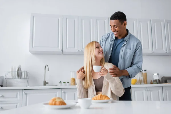 Afrikanisch-amerikanischer Mann lächelt junge blonde Freundin an, die mit Kaffeetasse neben verschwommenen Croissants sitzt — Stockfoto