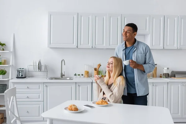 Alegre casal multiétnico com xícaras de café olhando para longe perto de deliciosos croissants na cozinha — Fotografia de Stock