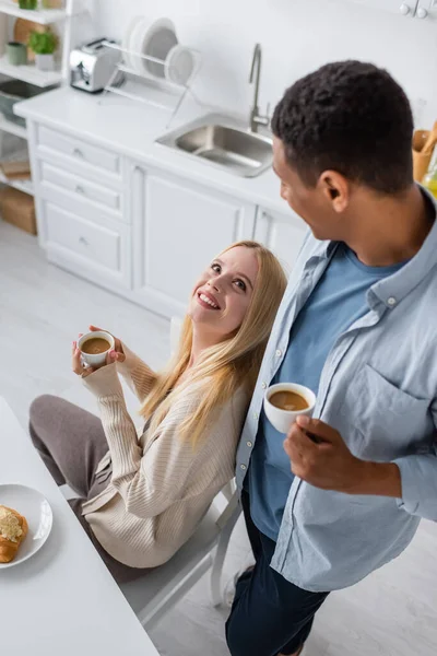 High angle view of blonde woman looking at african american boyfriend while drinking morning coffee in kitchen — Stock Photo