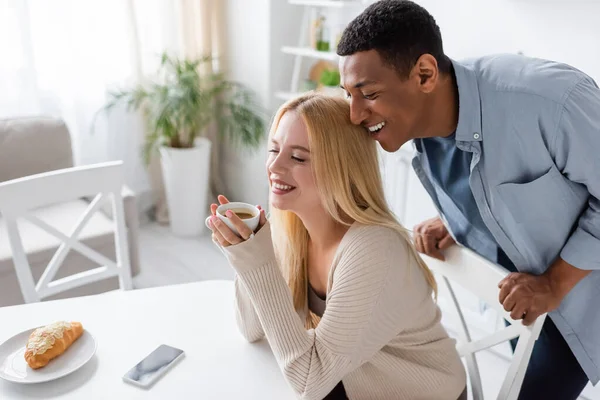 Веселый африканский американец, улыбающийся рядом с блондинкой, сидящей с чашкой кофе возле вкусного круассана — стоковое фото