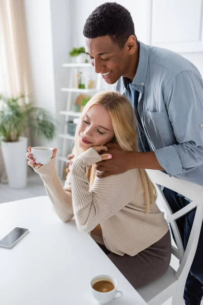 Afrikanisch-amerikanischer Mann umarmt junge Freundin sitzend mit geschlossenen Augen und hält Kaffeetasse — Stockfoto