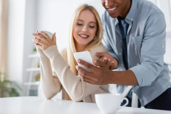 Sorrindo afro-americano homem mostrando celular para namorada borrada sentado com xícara de café na cozinha — Fotografia de Stock