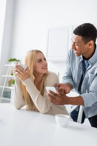 Cheerful african american man showing smartphone to blonde woman holding coffee cup — Stock Photo
