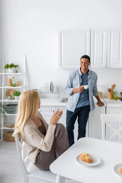 Lächelnder afrikanisch-amerikanischer Mann mit Kaffeetasse schaut blonde Freundin an, die neben Croissant auf dem Küchentisch sitzt — Stockfoto